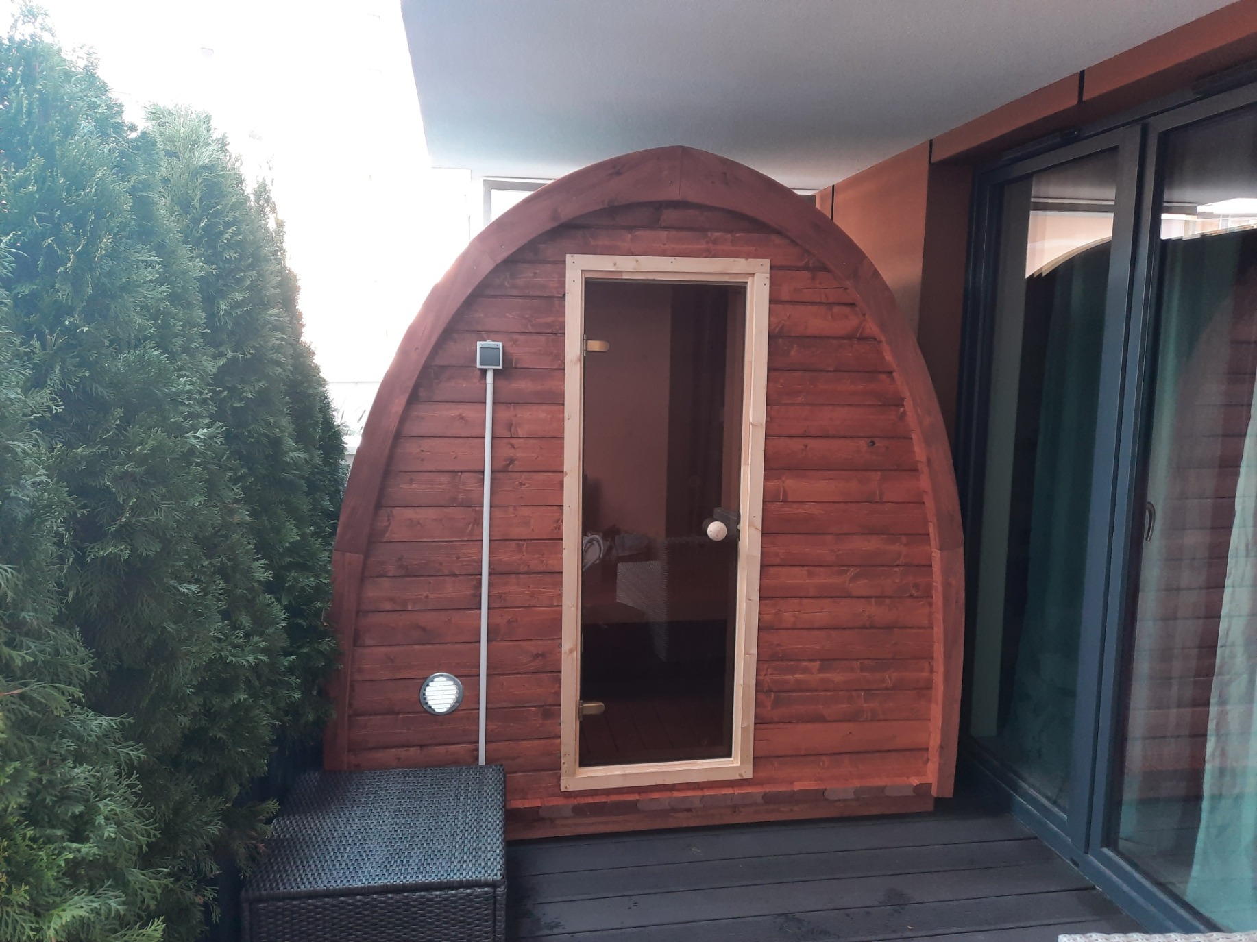 Drewniana sauna beczka z przeszklonymi drzwiami, ustawiona na balkonie obok tuj i mebli rattanowych.
