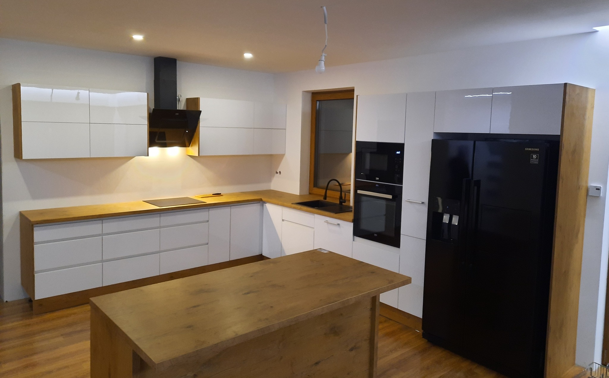 Nowoczesna kuchnia z białymi szafkami, drewnianym blatem i wyspą, czarnym sprzętem AGD oraz nieosłoniętą żarówką zwisającą z sufitu.