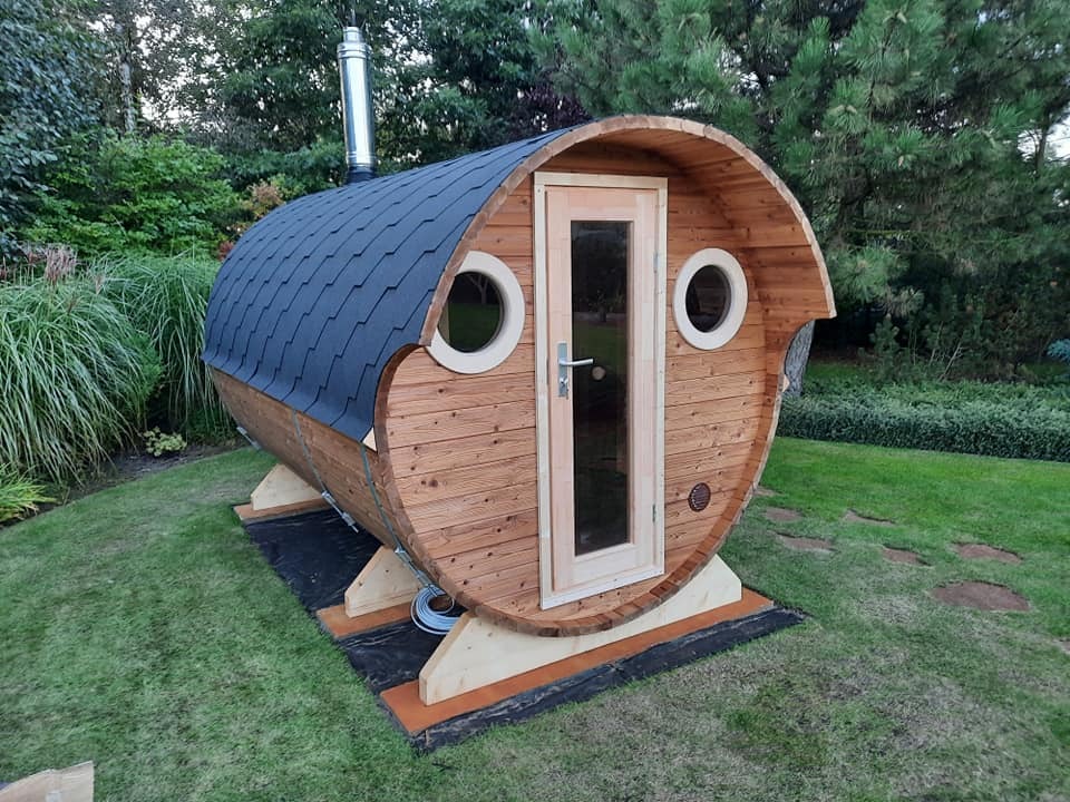 Drewniana sauna w kształcie beczki z oknami i kominem, ustawiona na trawie w ogrodzie.