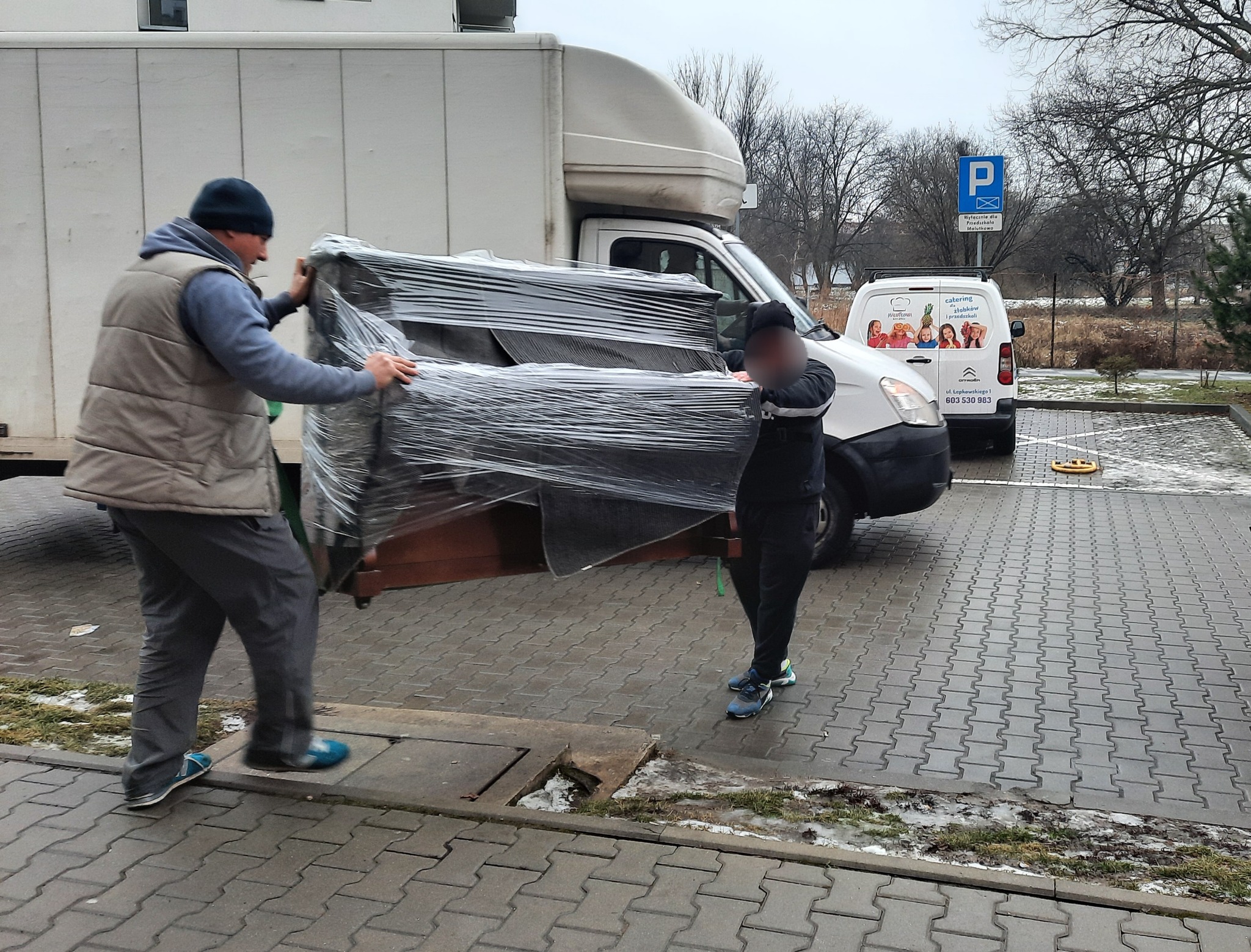 Dwóch mężczyzn w Krakowie przenosi owinięty folią mebel tapicerowany z chodnika do białej furgonetki, w tle zaparkowany samochód z reklamą cateringu dla dzieci.