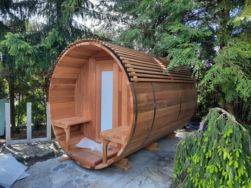 Drewniana beczka sauny ogrodowej z przeszklonymi drzwiami, umieszczona na betonowej powierzchni, otoczona zielenią drzew i krzewów.