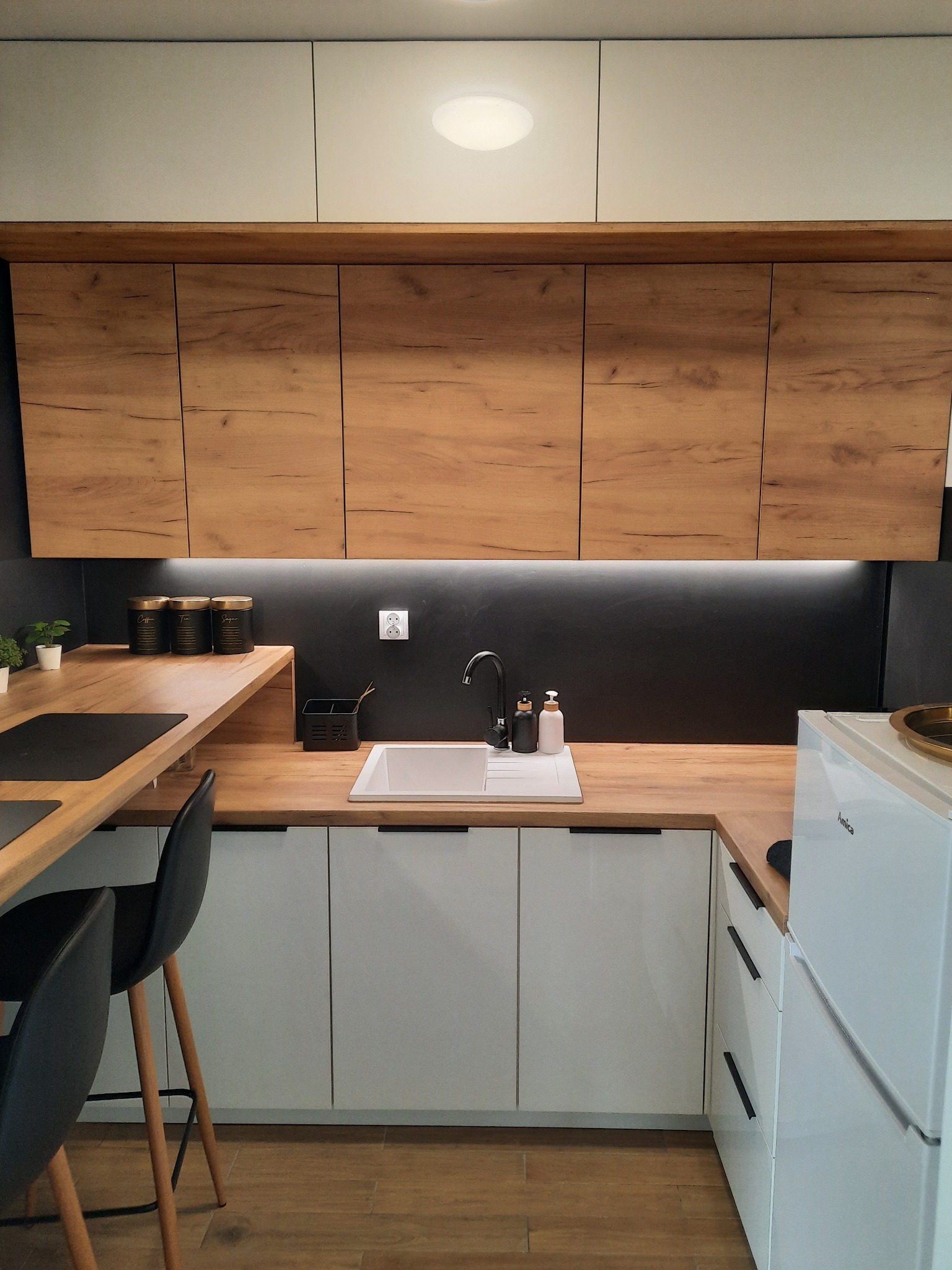 Nowoczesna kuchnia z drewnianymi i białymi szafkami, czarnym blatem i armaturą, oświetleniem LED pod szafkami wiszącymi i drewnianą podłogą.