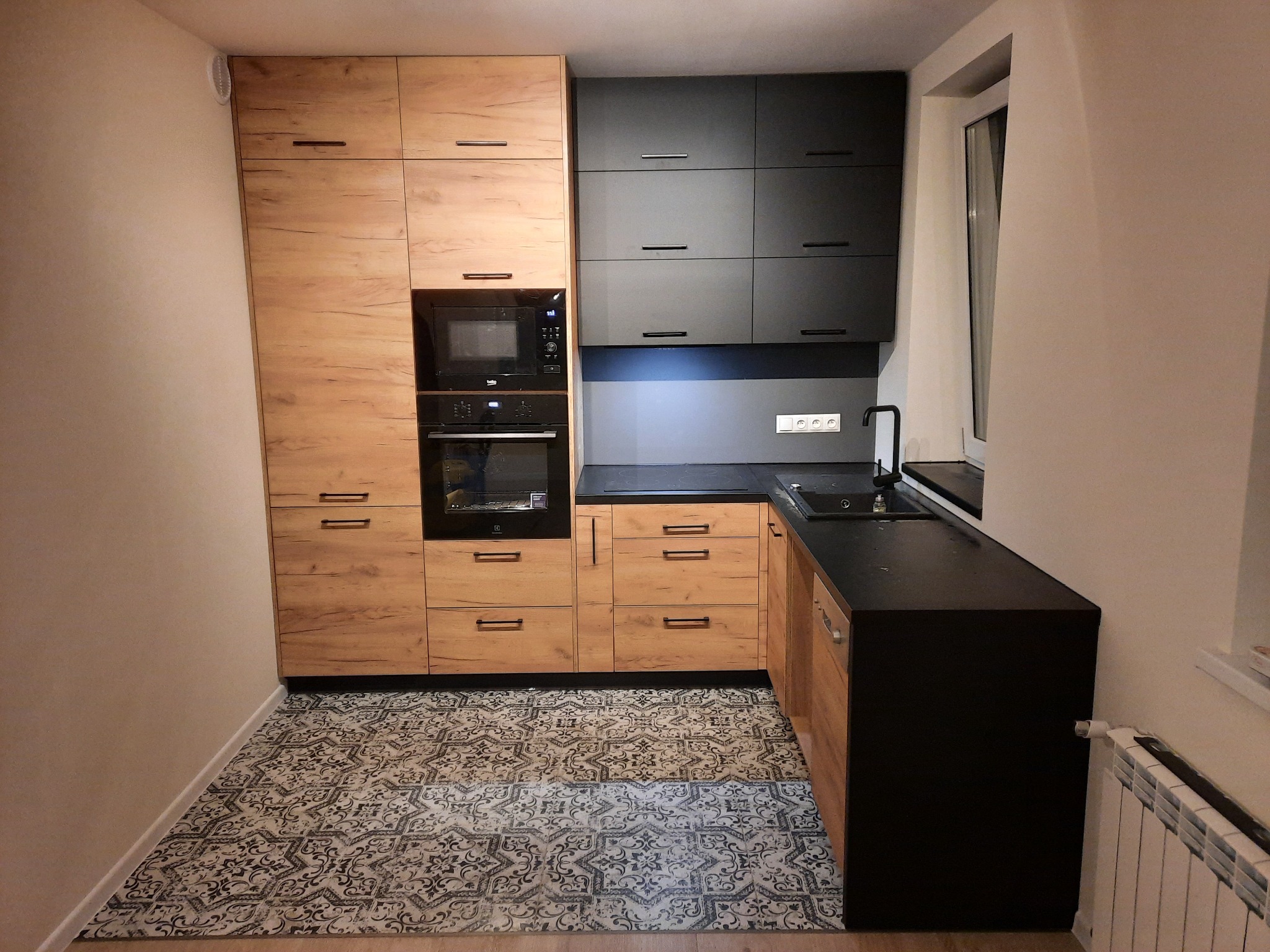 Nowoczesna kuchnia z drewnianymi szafkami z widoczną strukturą drewna, czarnymi szafkami wiszącymi, czarnym blatem i wzorzystą podłogą.