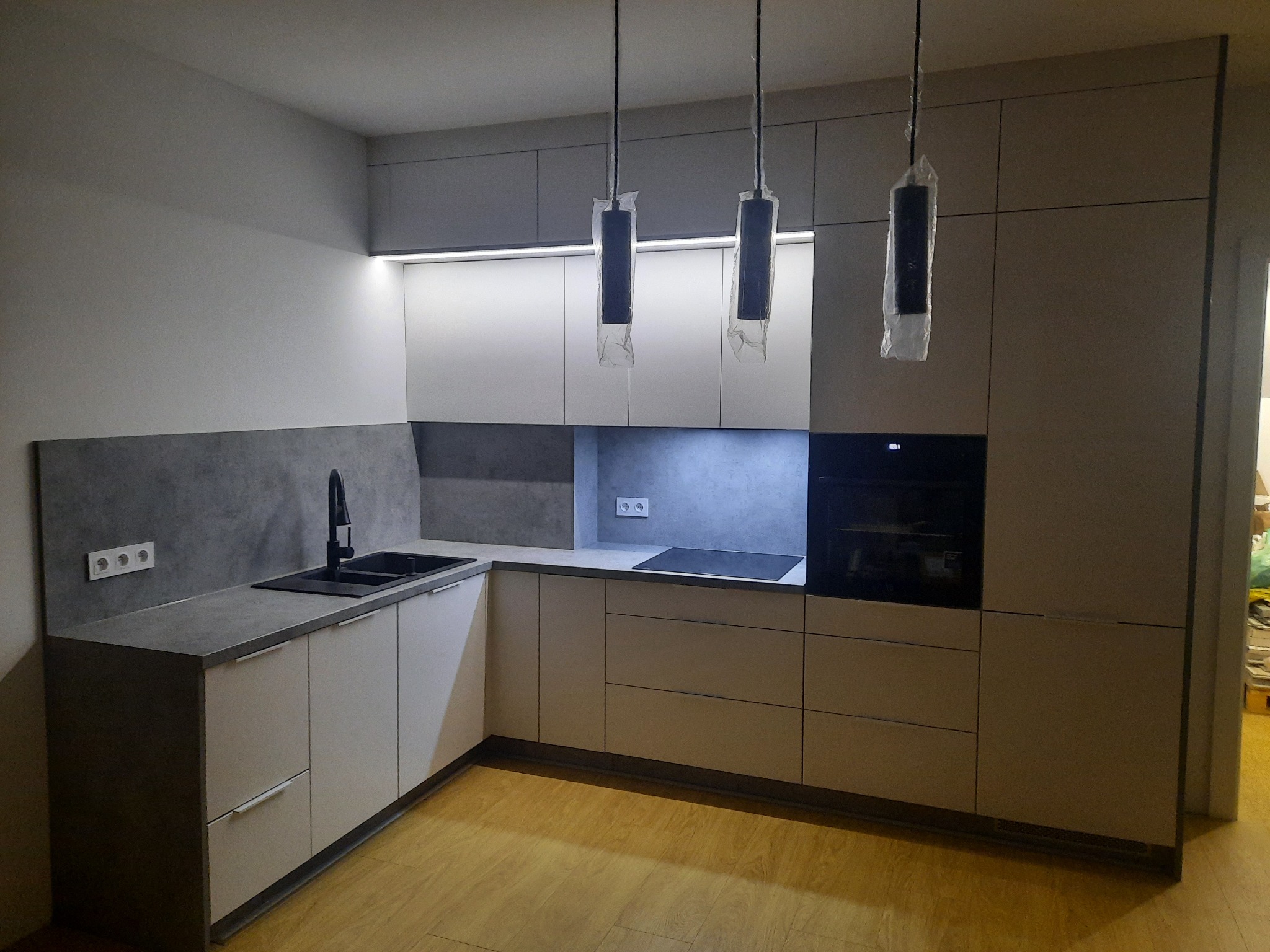 Nowoczesna kuchnia z szarymi i białymi szafkami, czarnym zlewem i baterią, oraz trzema lampami wiszącymi w plastikowych osłonach, oświetlona światłem LED pod szafkami.