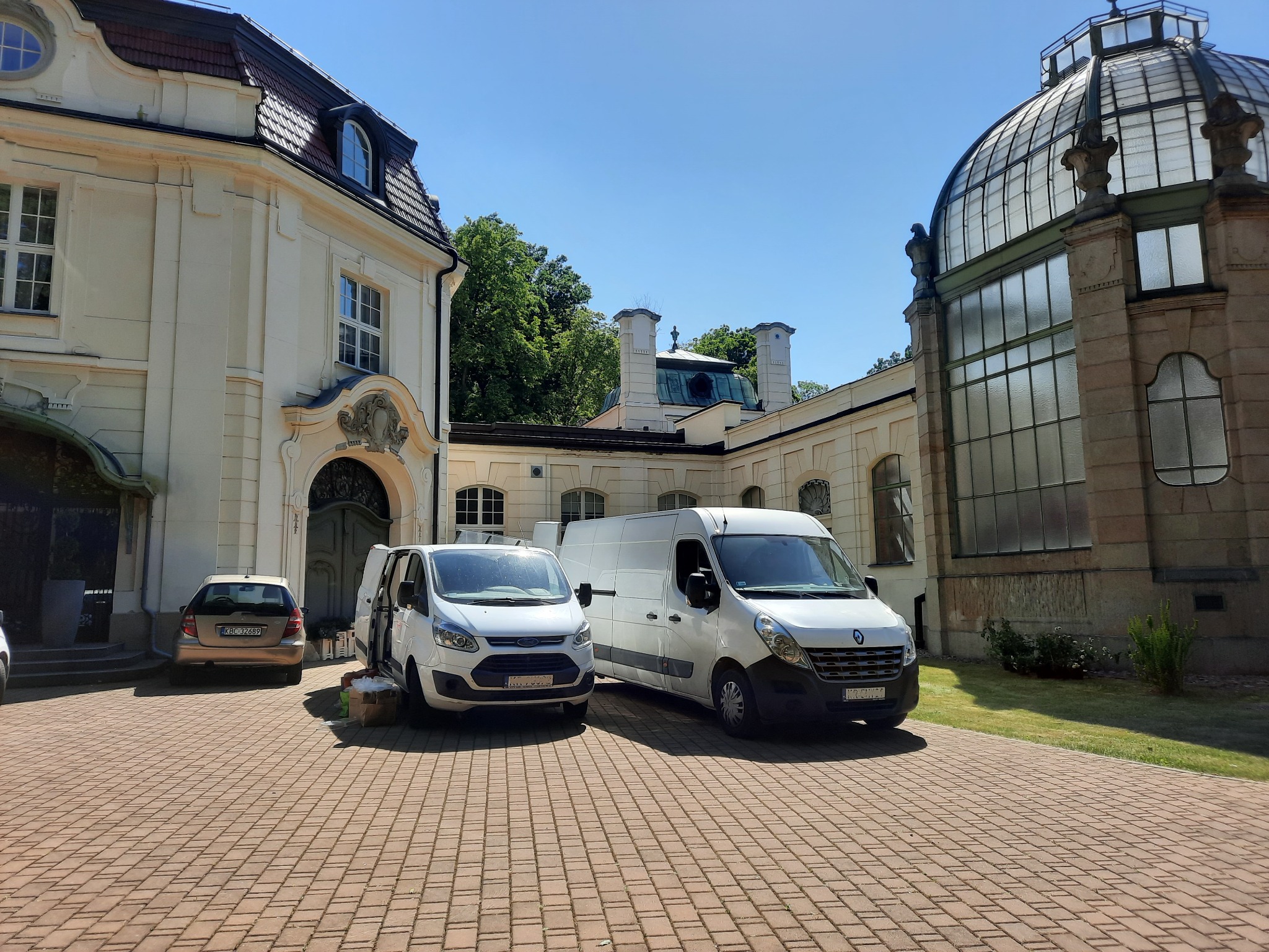 Dwa białe busy transportowe zaparkowane na brukowanym dziedzińcu przed zabytkowym budynkiem z oranżerią, sugerujące przeprowadzkę lub transport towarów.