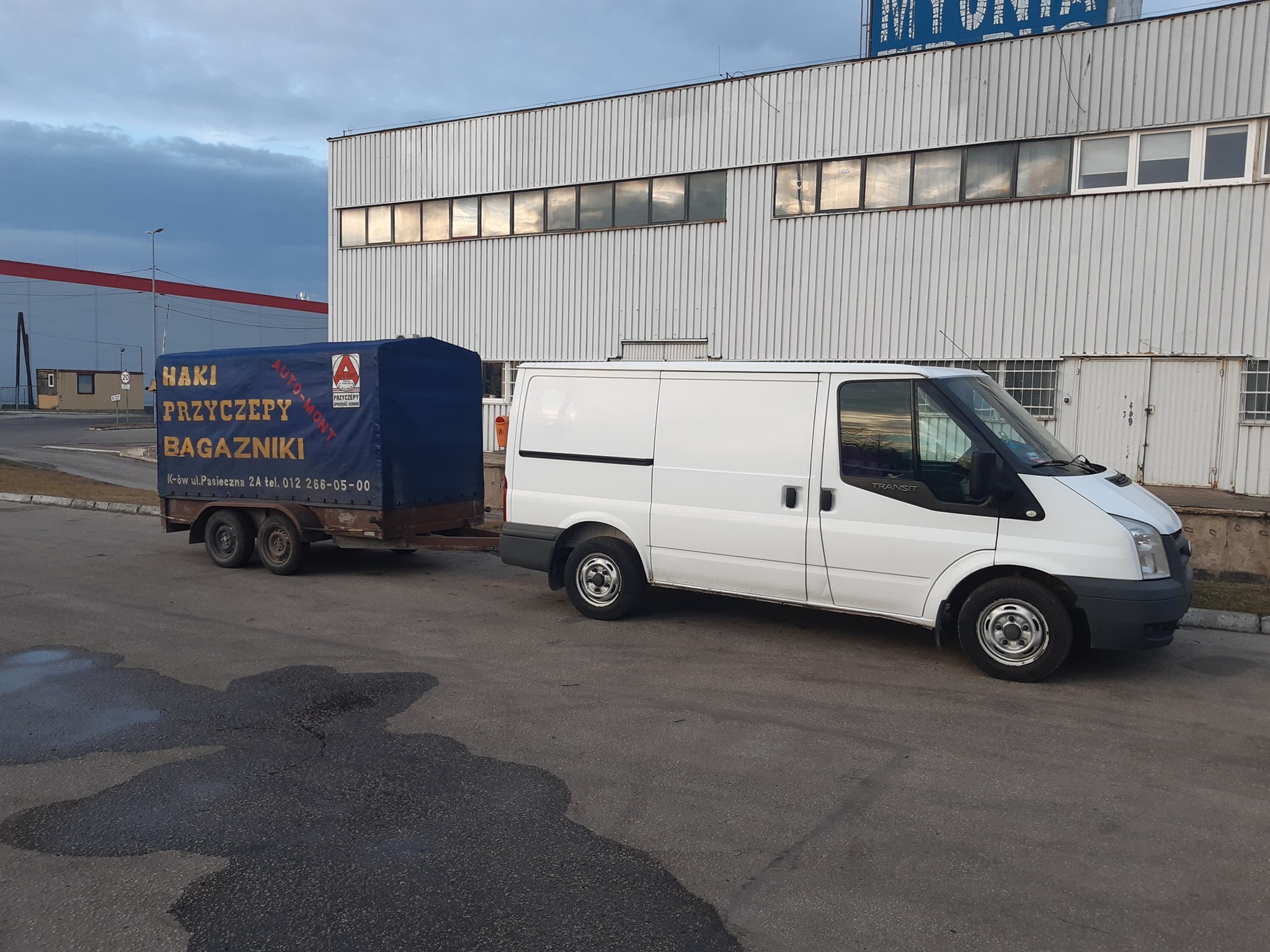 Biały samochód dostawczy Ford Transit z przyczepą, na której widnieje napis 'Haki Przyczepy Bagażniki', zaparkowany na tle hali przemysłowej z rzędem okien.