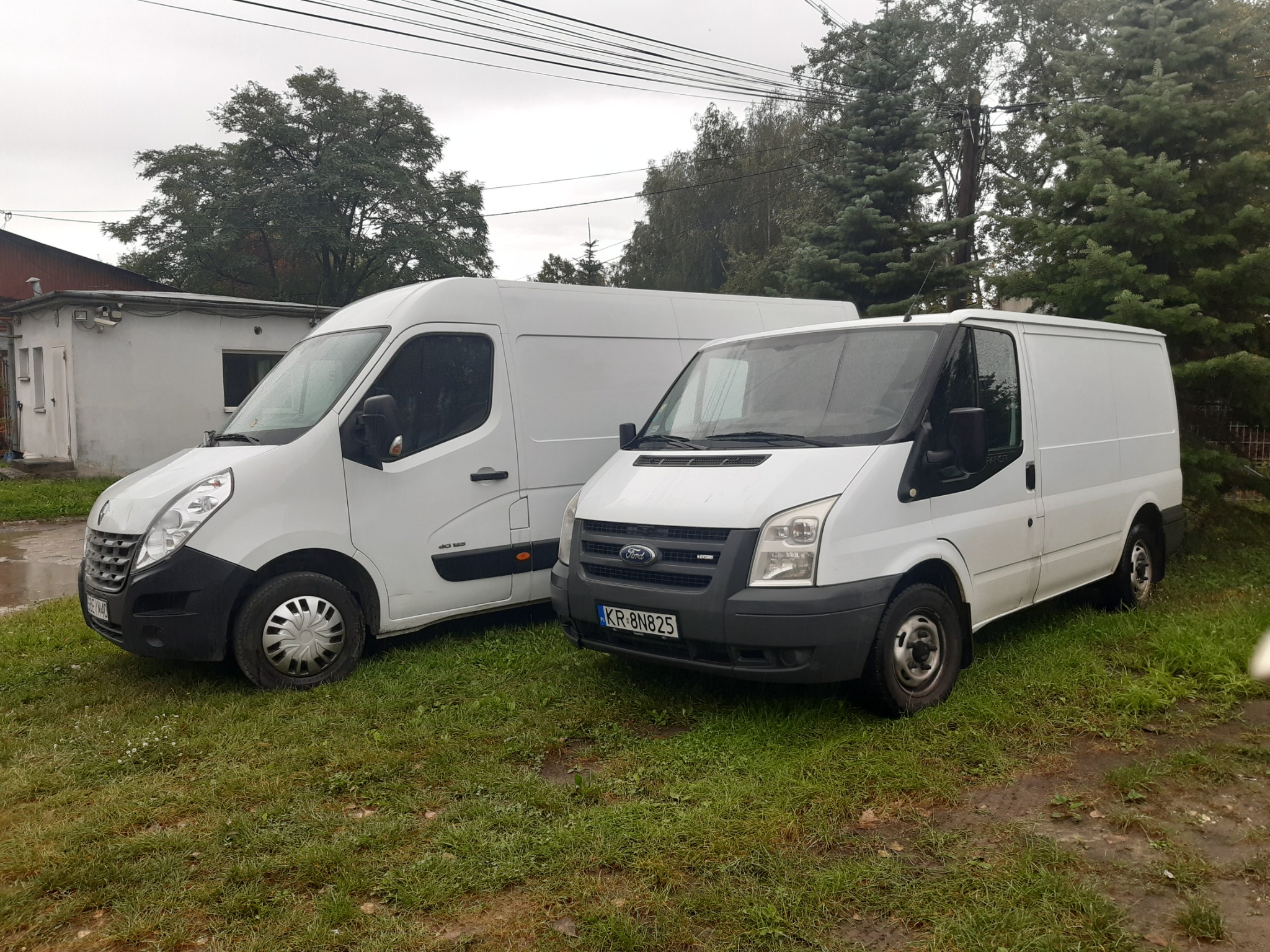 Dwa białe busy dostawcze, Renault Master i Ford Transit, zaparkowane na trawiastym terenie w pochmurny dzień. Ford Transit z widoczną tablicą rejestracyjną KR 8N825.