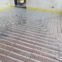 Montaż rur grzewczych na podłodze z siatką, widoczne żółte taśmy przy ścianach, surowy stan wykończenia.