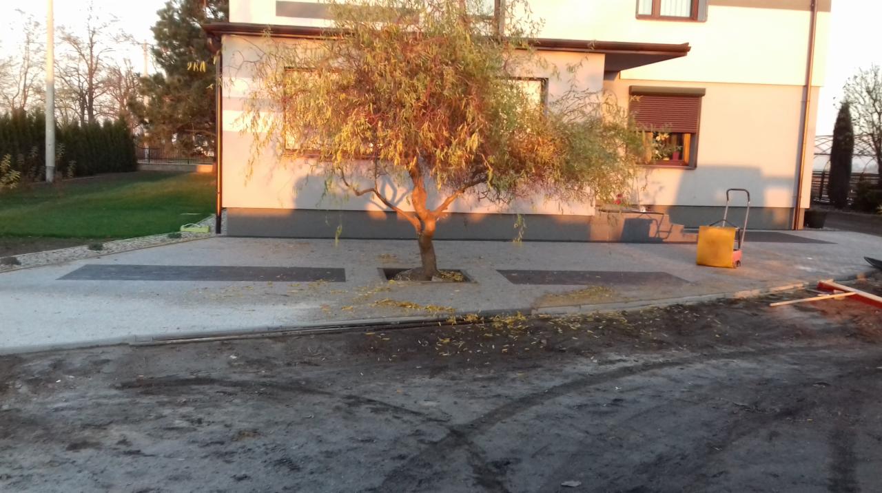 Ułożona nawierzchnia z kostki brukowej i płyt betonowych wokół drzewa, z widocznym fragmentem domu w tle i narzędziami budowlanymi po prawej stronie kadru.