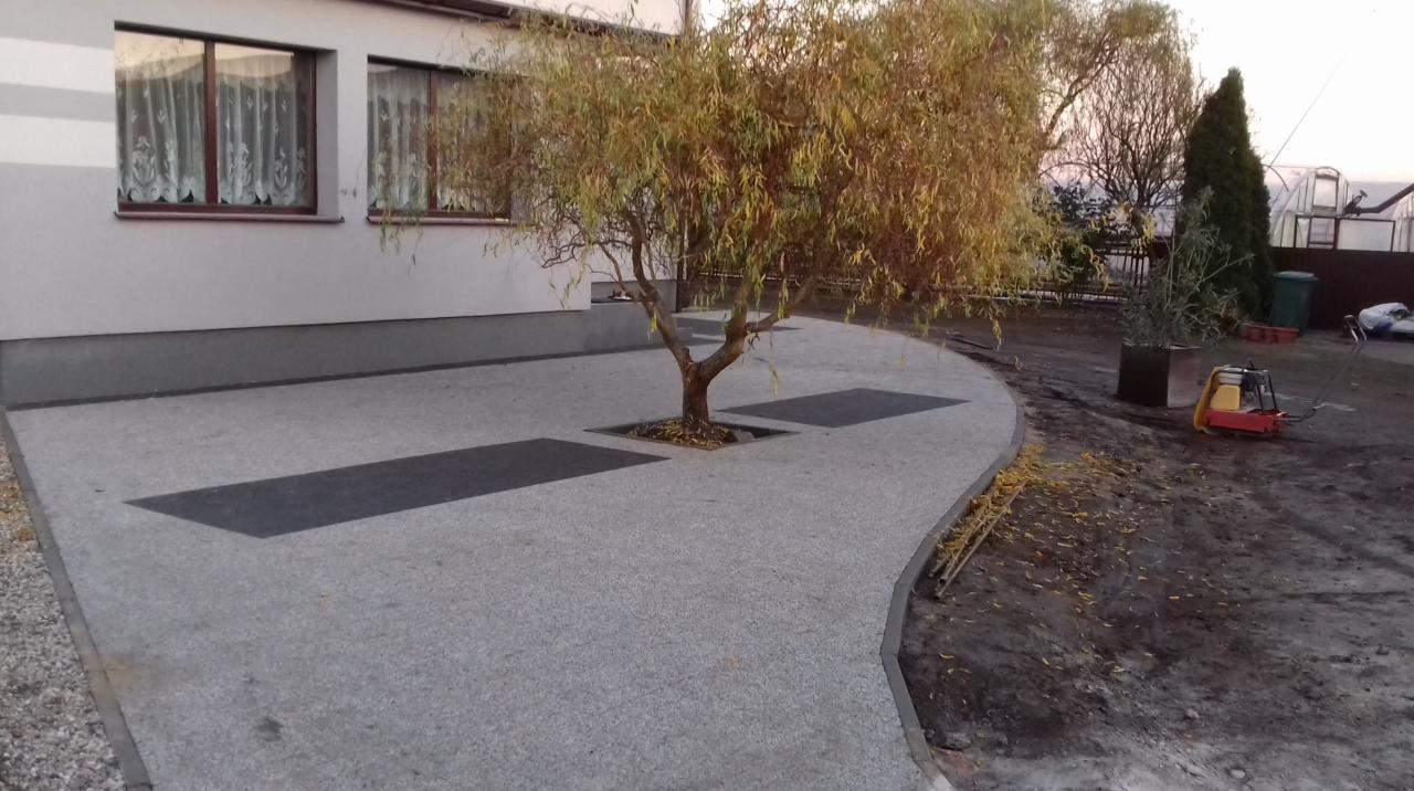 Szara nawierzchnia z kostki z wkomponowanym drzewem i ciemnymi prostokątnymi wstawkami, obok domu z oknami i zagęszczarką w tle.