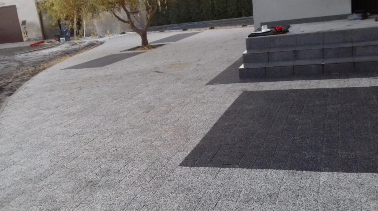 Ułożona kostka granitowa w odcieniach szarości prowadząca do schodów wejściowych, z wyznaczonym miejscem na drzewo, widoczny fragment placu budowy w tle.