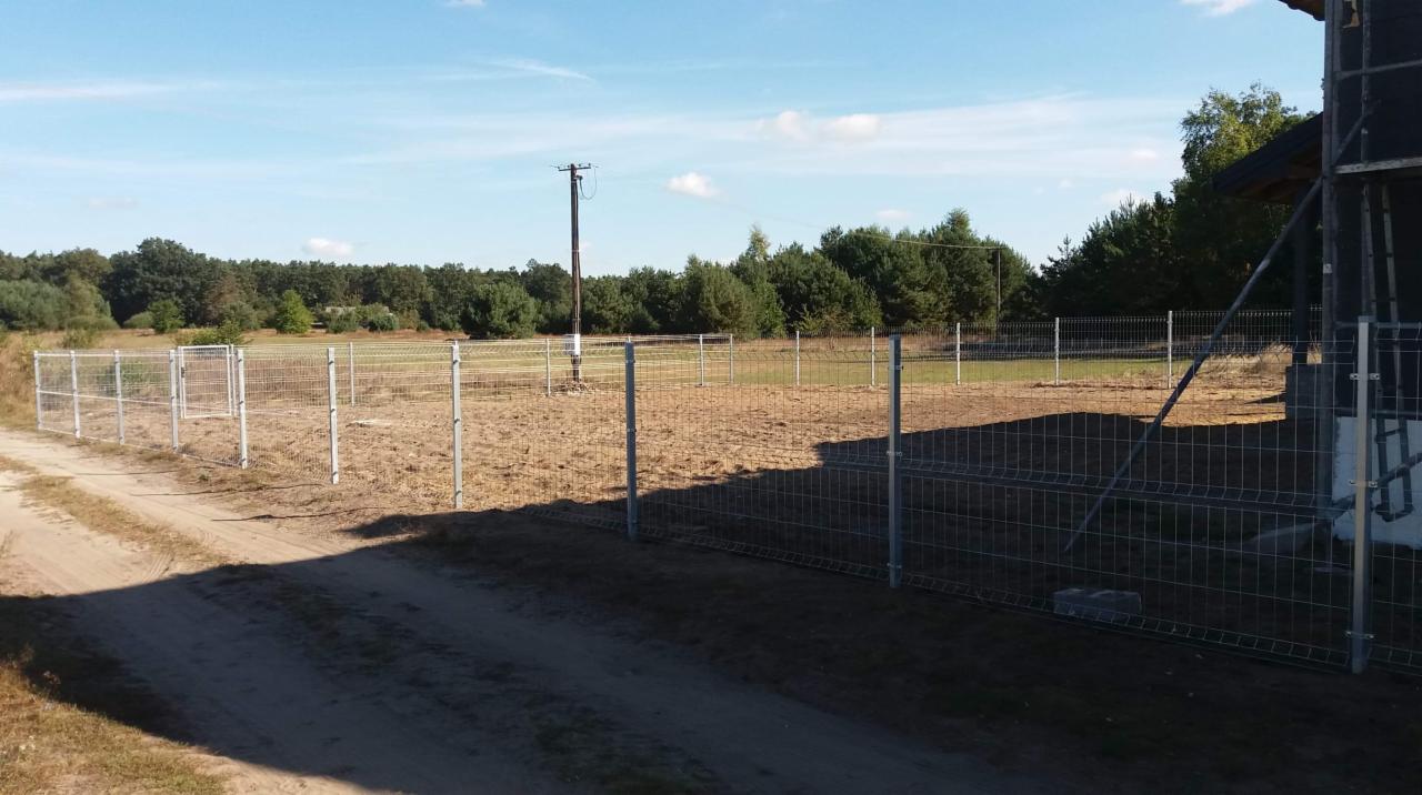 Metalowa siatka ogrodzeniowa z prostymi słupkami, otaczająca teren z suchą trawą, widoczny słup energetyczny i fragment domu w budowie.