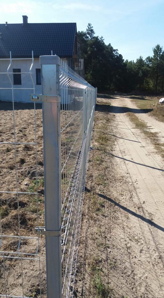 Metalowa siatka ogrodzeniowa zamontowana na posesji z widocznym domem w tle i piaszczystą drogą obok.