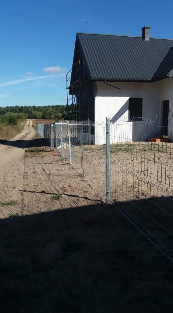 Nowoczesne ogrodzenie panelowe z drutu ocynkowanego, zabezpieczające posesję z nowym domem w tle, widoczne ślady opon na piaszczystym podłożu.