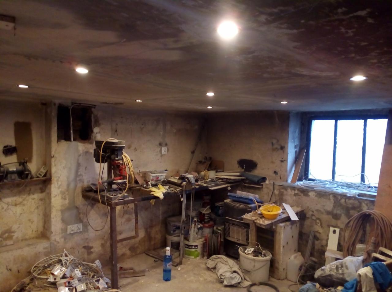 Piwnica w trakcie remontu z widoczną wiertarką stołową, puszkami farb, narzędziami i nowo zamontowanymi punktowymi źródłami światła w suficie.