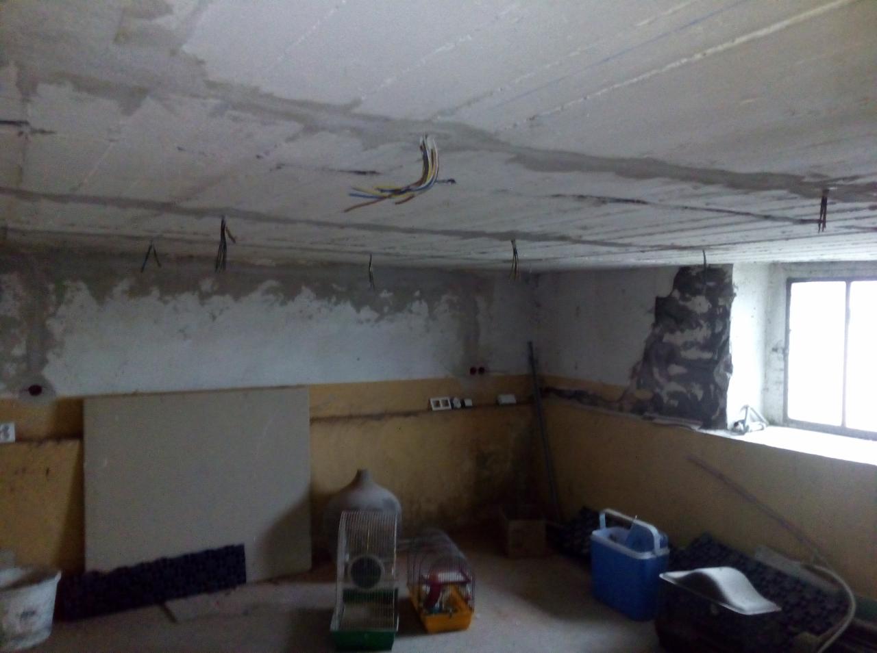 Surowe wnętrze w trakcie remontu: odsłonięty betonowy strop z wystającymi przewodami elektrycznymi, fragment ściany z widoczną cegłą, okno w tle i prowizoryczne przedmioty w pomieszczeniu.
