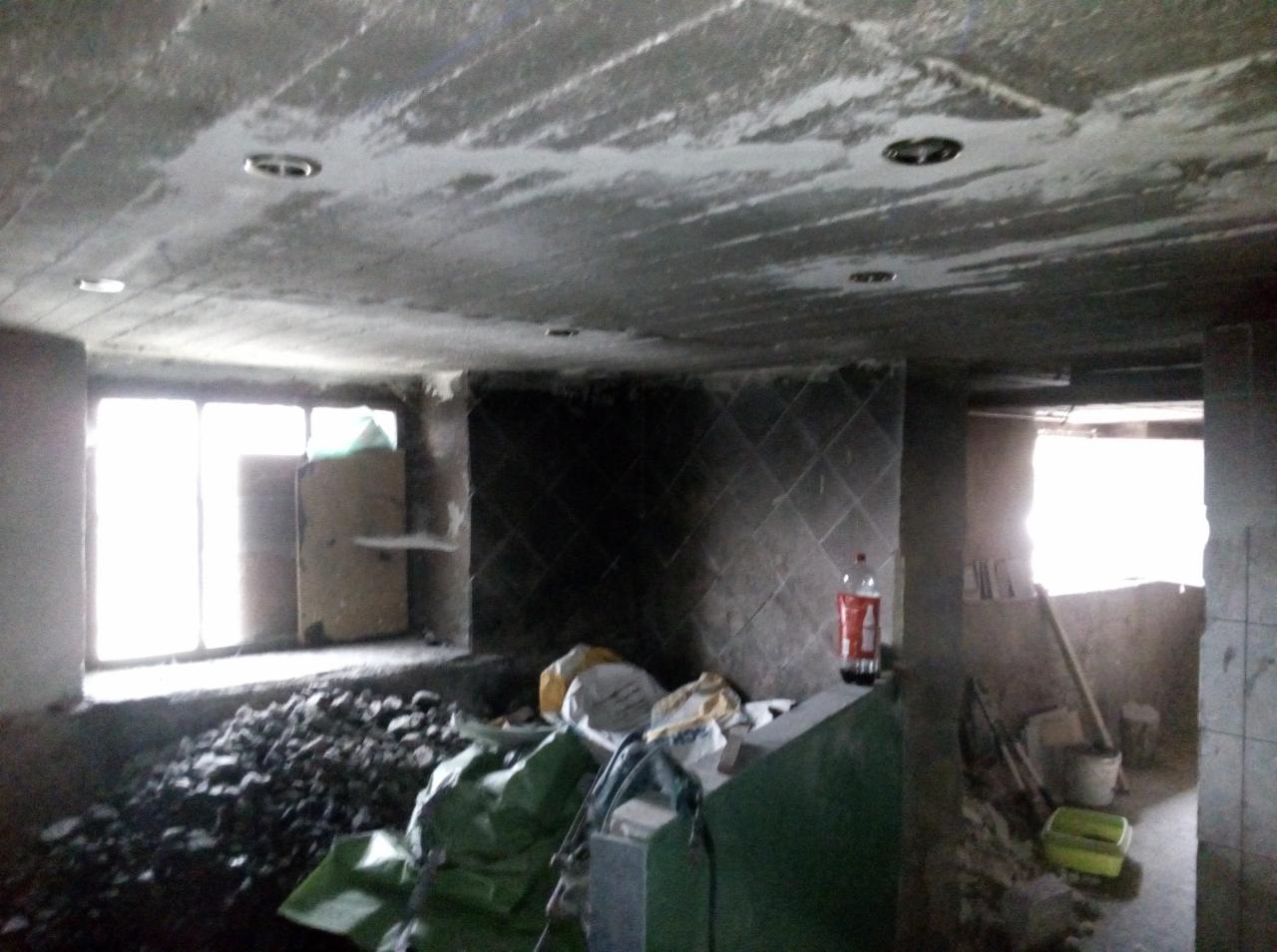 Surowy stan wnętrza w trakcie remontu: widoczne ślady po instalacji oświetlenia w suficie, gruz budowlany, worki z materiałami, częściowo wykończona ściana z płytek i butelka napoju.