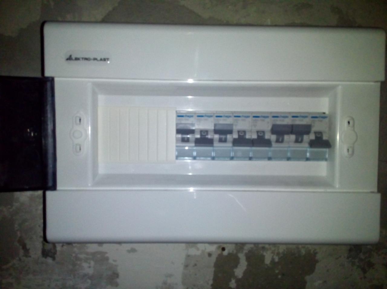 Biała skrzynka elektryczna firmy Elektro-Plast z otwartymi drzwiczkami, widoczne zabezpieczenia topikowe, zamontowana na szarej, surowej ścianie.