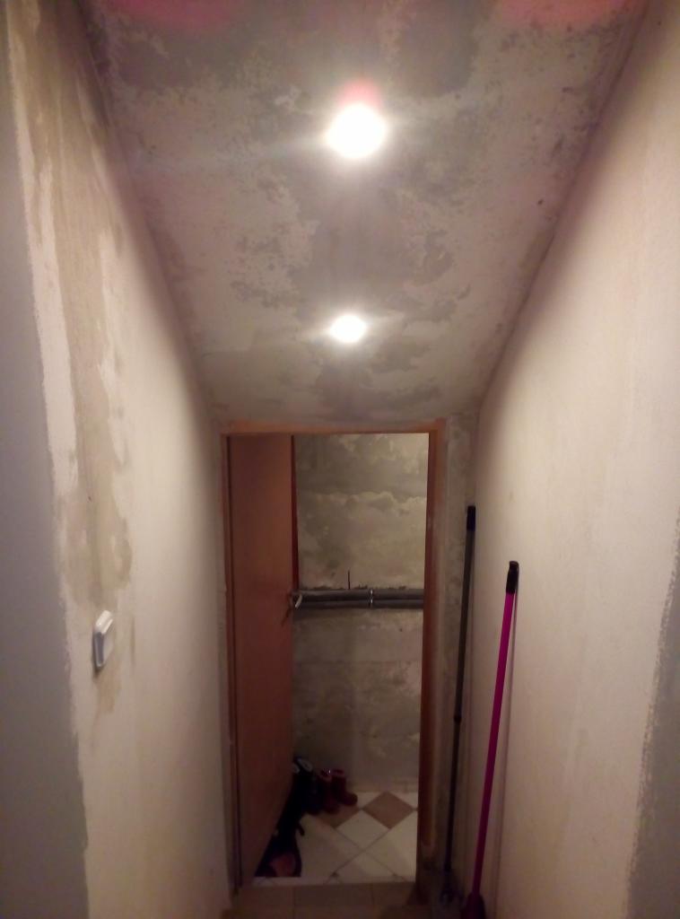 Korytarz w trakcie remontu z widocznymi śladami po malowaniu na ścianach i suficie, dwie lampy sufitowe, otwarte drzwi do pomieszczenia z rurami i narzędziami, oraz dwie miotły oparte o ścianę.