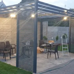 Nowoczesna, szara pergola z poziomymi listwami, oświetlona punktowymi lampami, z meblami ogrodowymi wewnątrz, na tle brukowanej nawierzchni i murowanego ogrodzenia.