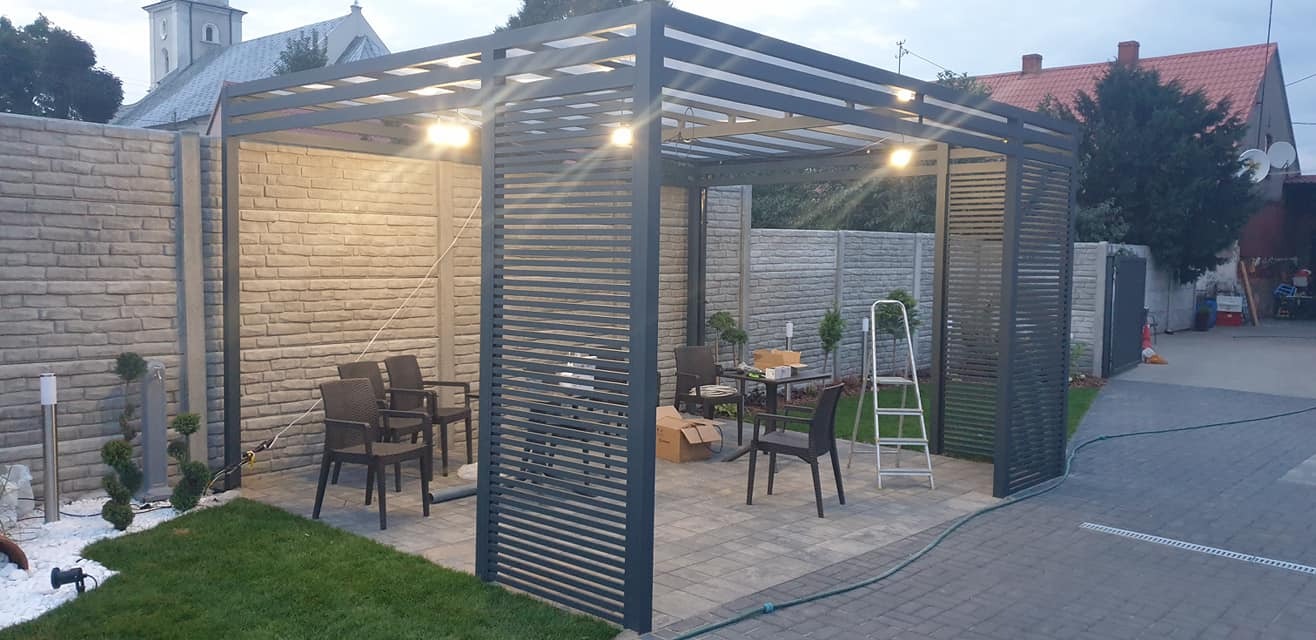 Nowoczesna, szara pergola z poziomymi listwami, oświetlona punktowymi lampami, z meblami ogrodowymi wewnątrz, na tle brukowanej nawierzchni i murowanego ogrodzenia.