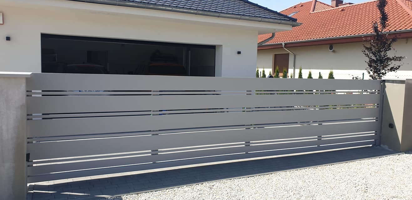 Nowoczesna, szara, przesuwna brama posesyjna z poziomymi panelami, widok z zewnątrz na tle białego domu z garażem i czerwonym dachem sąsiedniego budynku.