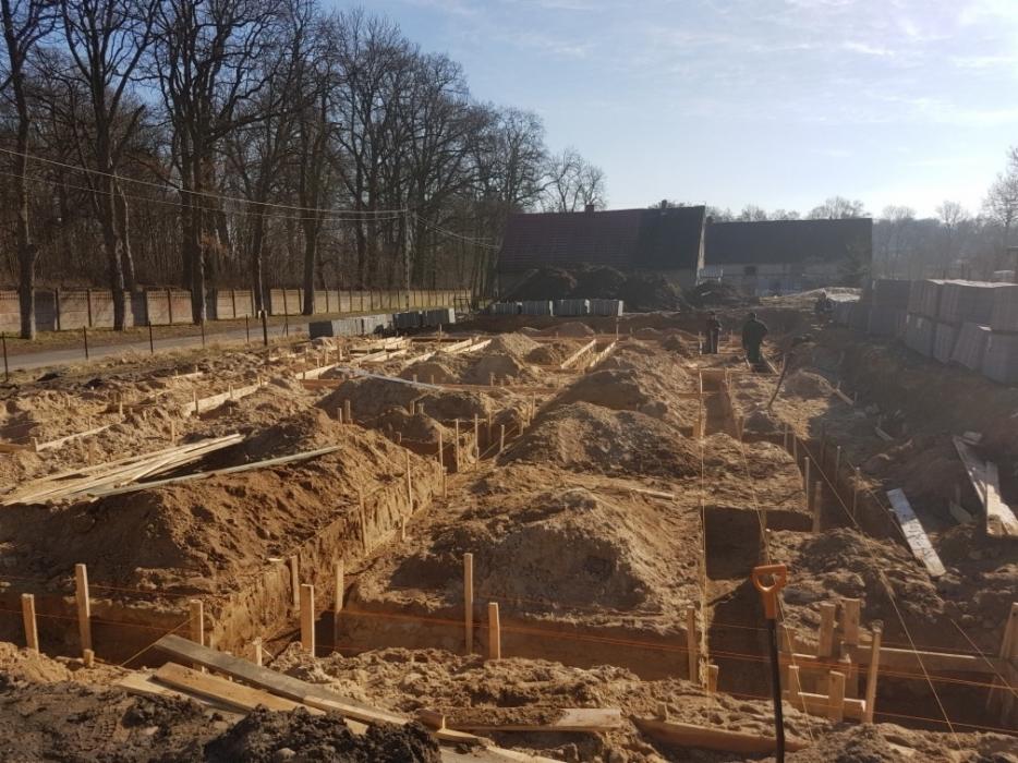 Przygotowanie fundamentów pod budowę domu: wykopane rowy z drewnianym szalunkiem, widoczny stos bloczków fundamentowych, ziemia i narzędzia.