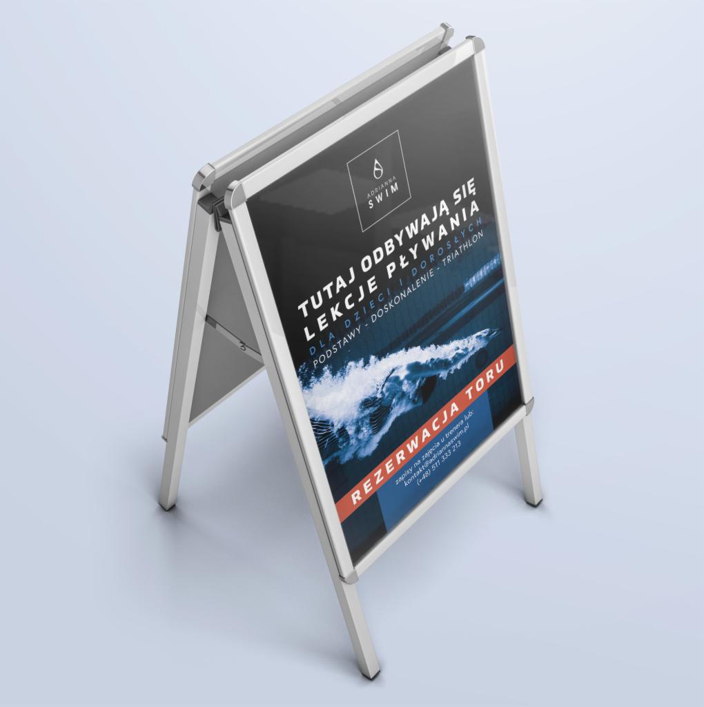Stojak reklamowy z plakatem informującym o lekcjach pływania dla dzieci i dorosłych, z logo Adrianna Swim i informacją o rezerwacji toru.
