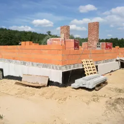 Wznoszenie ścian domu z bloczków ceramicznych Porotherm, widoczne fundamenty, izolacja przeciwwilgociowa, palety z bloczkami i kominy w trakcie budowy na tle lasu i błękitnego nieba.