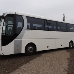 F.H.U. Wolbus - Biały autokar turystyczny z czarnymi szybami i szarymi akcentami, zaparkowany na żwirowym parkingu w pochmurny dzień.