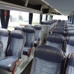 F.H.U. Wolbus - Wnętrze autobusu z rzędami niebieskich foteli pasażerskich z zagłówkami i pasami bezpieczeństwa, widok przez okno na inne pojazdy w deszczowy dzień.