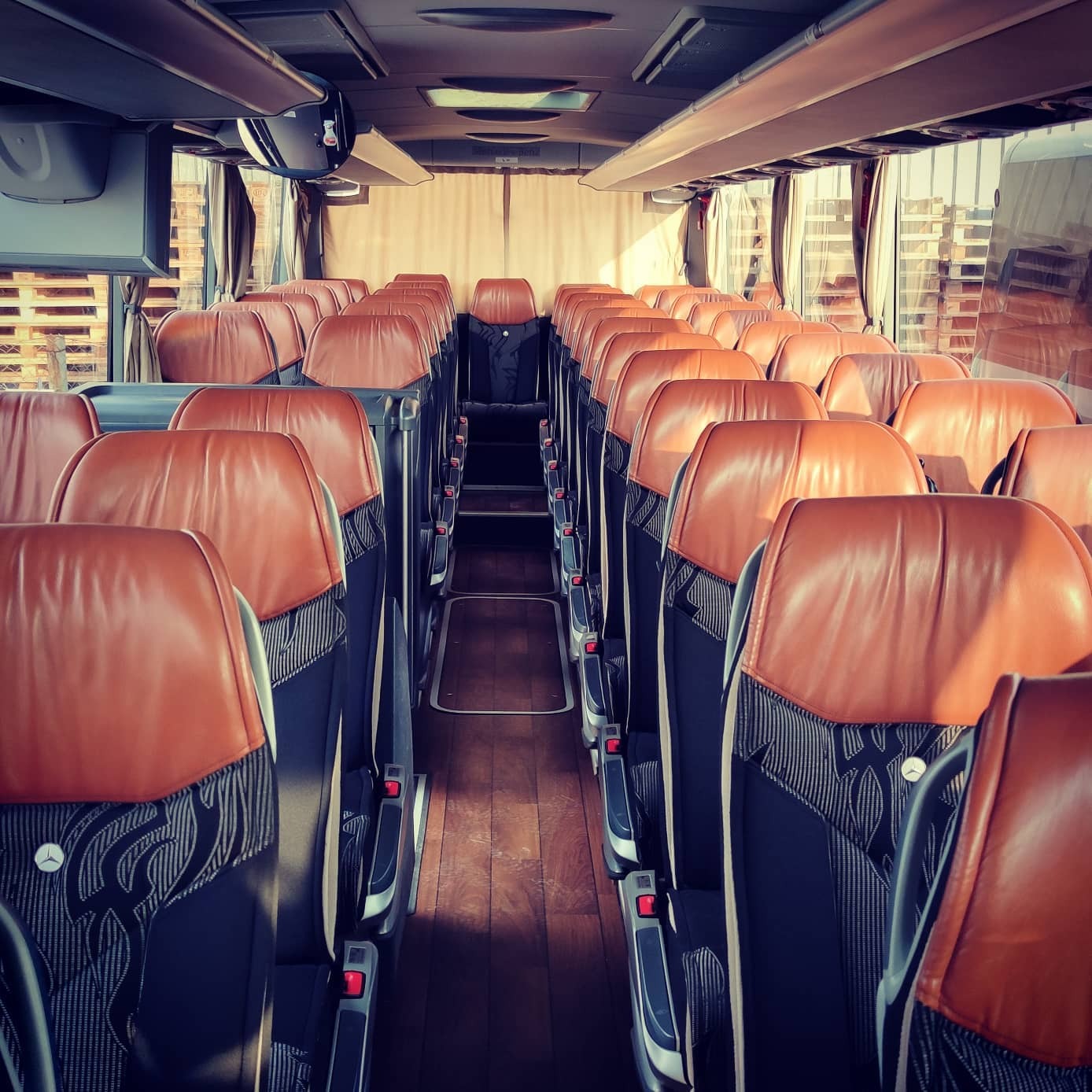 Wnętrze autobusu turystycznego z brązowymi skórzanymi zagłówkami foteli i wzorzystą tapicerką, widok na puste przejście i tylne okno z widocznymi paletami na zewnątrz.