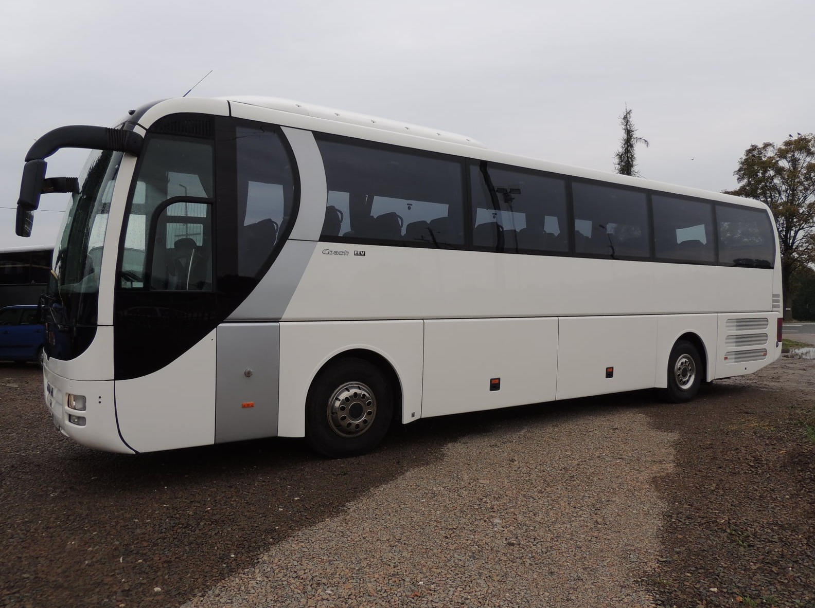 Biały autokar turystyczny z czarnymi szybami i szarymi akcentami, zaparkowany na żwirowym parkingu w pochmurny dzień.