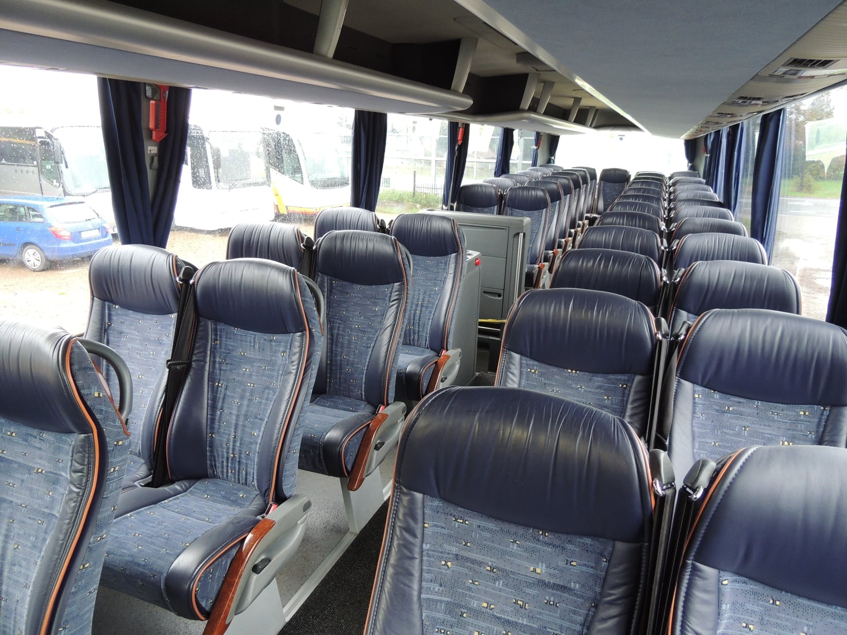 Wnętrze autobusu z rzędami niebieskich foteli pasażerskich z zagłówkami i pasami bezpieczeństwa, widok przez okno na inne pojazdy w deszczowy dzień.