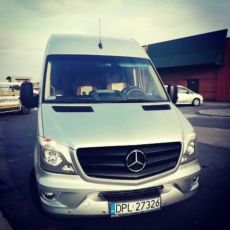 Srebrny bus Mercedes-Benz Sprinter z widoczną tablicą rejestracyjną DPL 27326, zaparkowany na parkingu przed budynkiem z zielonym dachem.
