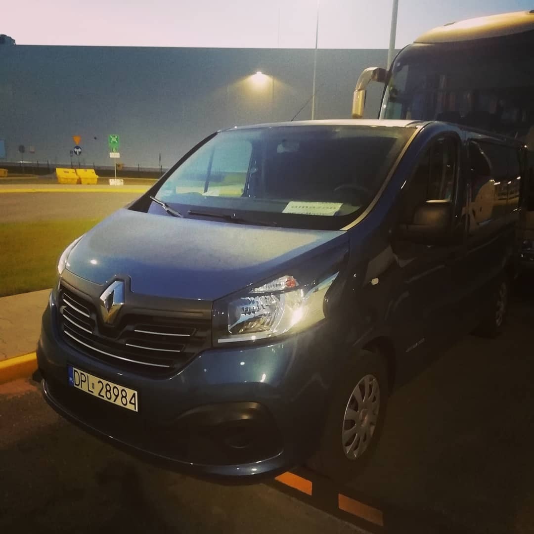 Niebieski bus Renault Trafic z widoczną tablicą rejestracyjną DPL 28984, zaparkowany na tle szarego budynku magazynowego i autobusu, oświetlony wieczornym światłem.