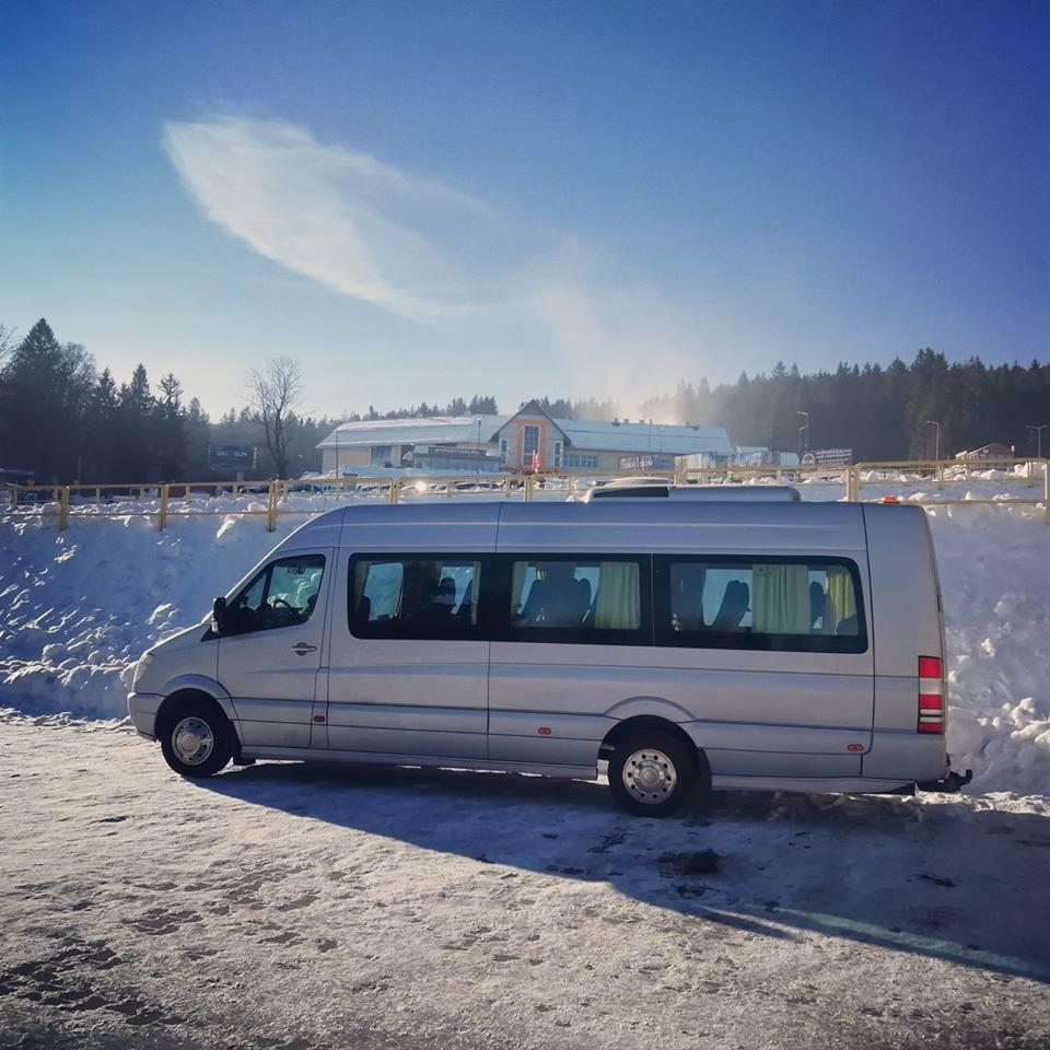 Srebrny bus pasażerski zaparkowany na ośnieżonym parkingu przed budynkiem w zimowej scenerii, widoczne odbicia w szybach.