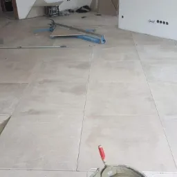 Układanie jasnych płytek podłogowych w dużym pomieszczeniu, widoczny fragment narzędzia do cięcia płytek i wiadro z zaprawą.