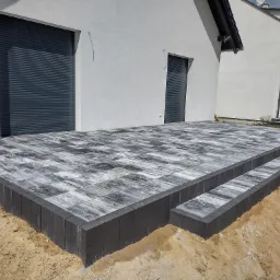 Nowoczesny taras z kostki brukowej w odcieniach szarości, z dwustopniowymi schodami, na tle białego budynku z szarymi roletami.