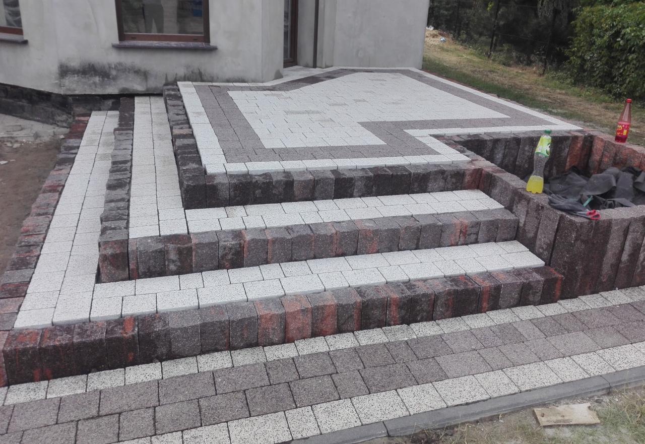 Schody wejściowe do domu wyłożone kostką brukową w odcieniach szarości i czerwieni, z donicą z kostki po prawej stronie, butelka wody i napoju w tle.