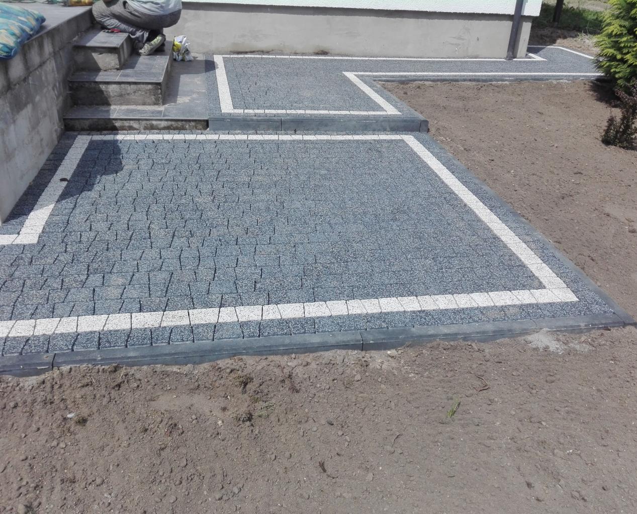 Szara kostka brukowa ułożona w geometryczny wzór z białym obramowaniem, widoczne schody wejściowe do budynku i fragment trawnika.