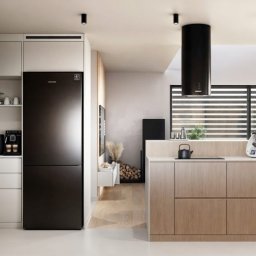 Aleksandra Lachman - Nowoczesna kuchnia w jasnych barwach z czarną lodówką i okapem. Minimalistyczny design, drewniane wykończenia, oświetlenie punktowe i duża powierzchnia blatu roboczego.