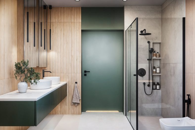 Nowoczesna łazienka z drewnianą ścianą, podwójną umywalką i zielonymi akcentami kolorystycznymi w postaci szafki i drzwi. Kabina prysznicowa ze szklanymi ścianami.