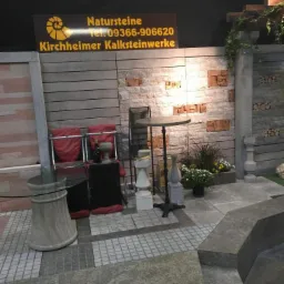 Ekspozycja kostki brukowej i granitowej z elementami dekoracyjnymi: stoły, krzesła, rośliny, fragmenty ścian z różnego rodzaju kamienia, oraz szyld reklamowy firmy Kirchheimer Kalksteinwerke.