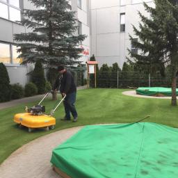 PPUH Nastała Lucjan - Mężczyzna w czapce kosi żółtą kosiarką trawnik przed budynkiem, w tle widoczne zielone pokrowce na piaskownicach i drzewa iglaste.