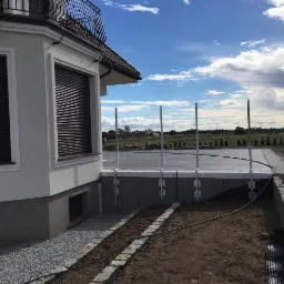 Przygotowanie podłoża pod taras z widocznymi elementami konstrukcyjnymi i instalacją nawadniającą w ogrodzie, na tle błękitnego nieba z chmurami.