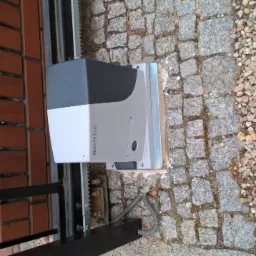 Szary automat do bramy Nice Robus zamontowany na metalowej bramie wjazdowej, widok z boku na tle muru z czerwonej cegły i kostki brukowej.