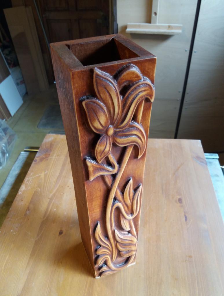 Wysoka, kwadratowa, drewniana donica z rzeźbionym motywem kwiatowym, stojąca na drewnianym stole.