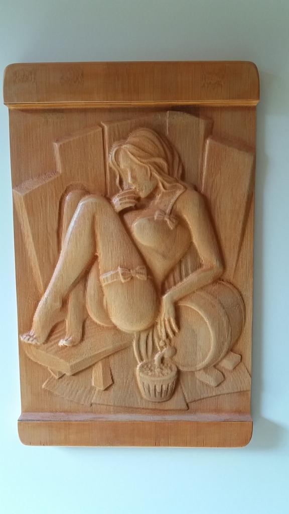 Drewniana płaskorzeźba przedstawiająca siedzącą kobietę w bieliźnie z kokardkami, trzymającą filiżankę, w tle abstrakcyjne kształty przypominające meble.