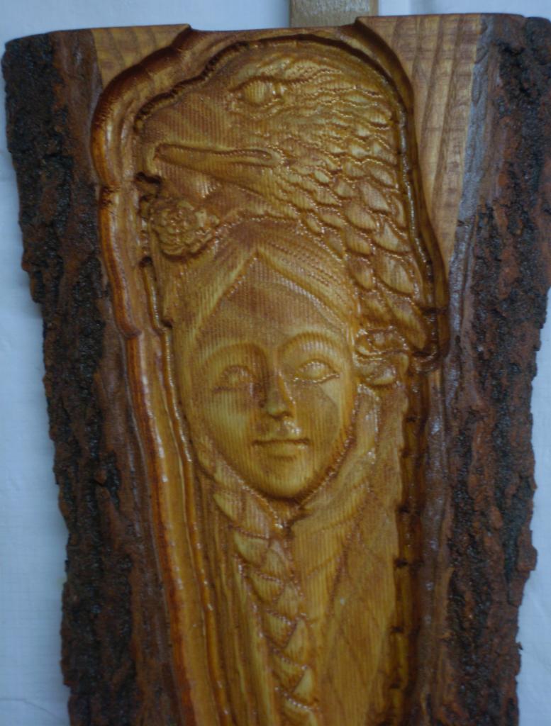 Ręcznie rzeźbiona płaskorzeźba w drewnie przedstawiająca twarz kobiety z warkoczem i orłem nad nią, osadzona w naturalnej korze drzewa.